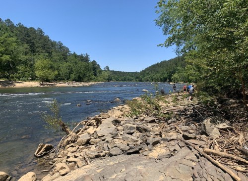 A scenic riverbank with rocky terrain, surrounded by lush greenery and people walking along the water's edge.