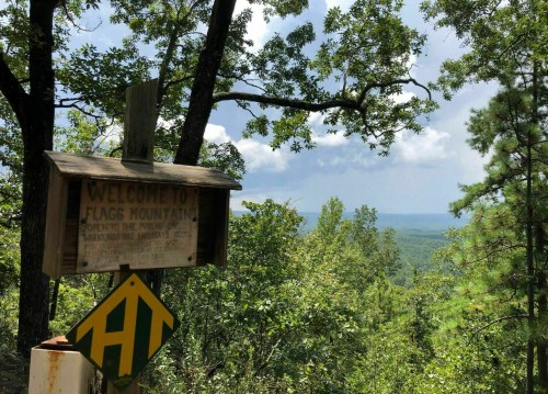 A wooden sign welcomes visitors to Flag Mountain, surrounded by lush trees and a scenic view of rolling hills.