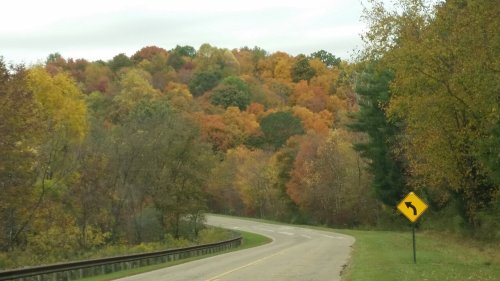 A winding road curves through a vibrant autumn landscape with colorful trees and a caution sign.