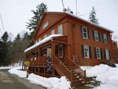 A large orange house with a sloped roof, surrounded by snow, featuring a porch and multiple windows.