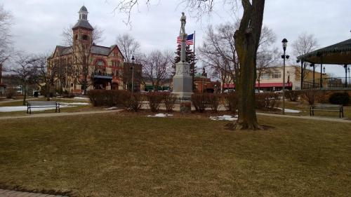 A park with a monument, flags, and a historic building in the background, surrounded by bare trees and grass.