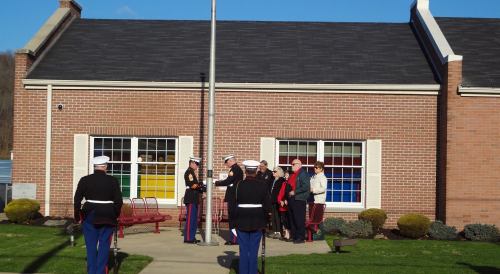A group gathers near a flagpole as Marines conduct a ceremony outside a brick building on a sunny day.