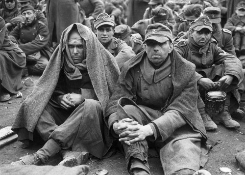 Black and white photo of weary soldiers sitting together, wrapped in blankets, with expressions of fatigue and resilience.