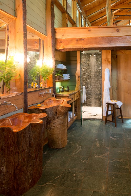 A rustic bathroom with wooden sinks, stone flooring, and a glass shower, featuring natural light and greenery.