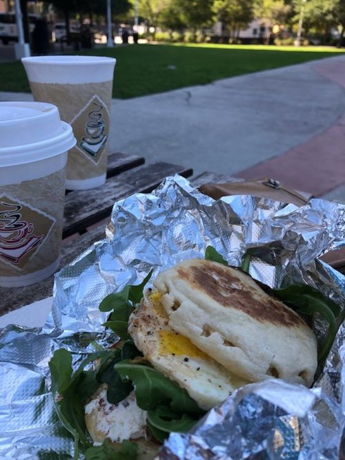 A close-up of a sandwich with egg and greens wrapped in foil, with two coffee cups in the background on a park bench.