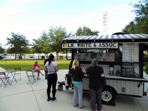 A food cart named "Yolk White & Assoc" with customers ordering, set in a park with trees and a lake in the background.