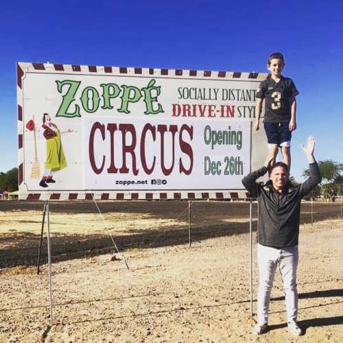 A boy stands on a man's shoulders in front of a sign for Zoppé Circus, promoting a drive-in event opening December 26th.
