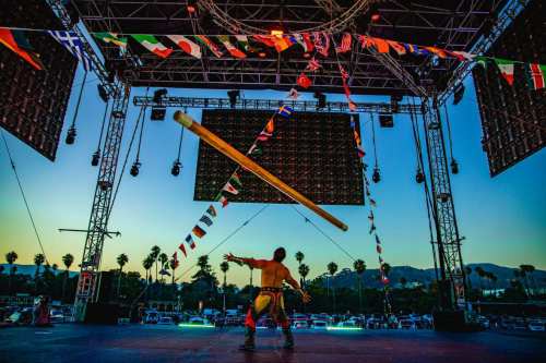 A performer on stage spins a large pole, surrounded by colorful flags and a vibrant sunset backdrop.