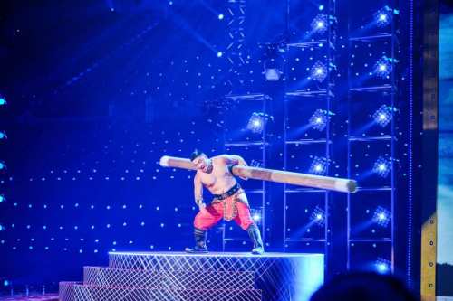 A performer in red pants lifts a large wooden pole on stage, surrounded by bright blue lights and a cheering audience.