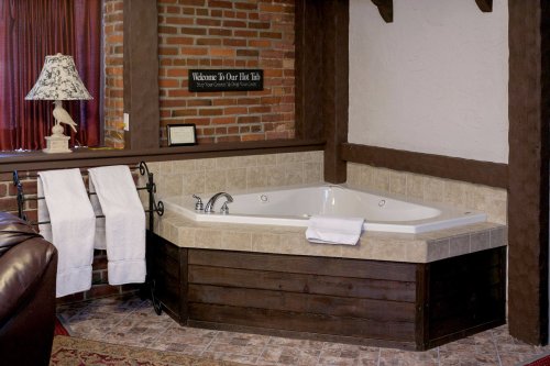A cozy bathroom featuring a corner jacuzzi tub, towels, a lamp, and rustic brick walls.