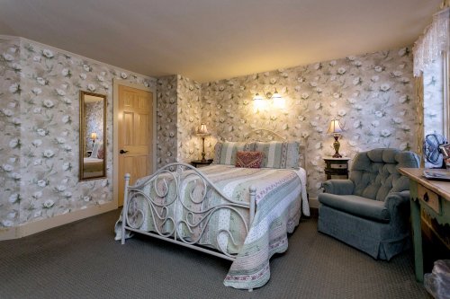Cozy bedroom with floral wallpaper, a bed with a quilt, a mirror, two lamps, and a blue armchair.
