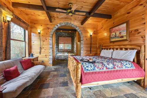 Cozy wooden bedroom with a stone fireplace, quilted bedspread, and a sofa, surrounded by large windows.