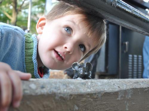 A young boy with blue eyes curiously looks at a small mouse figurine on a concrete surface.