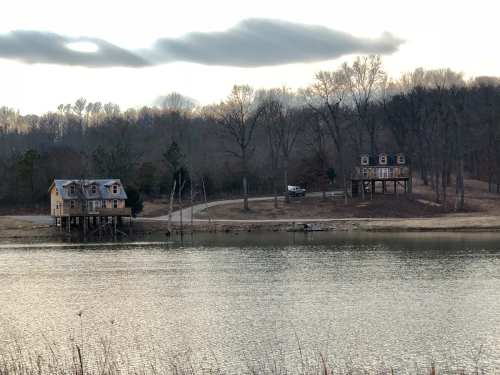 Two houses on stilts by a calm lake, surrounded by trees and a cloudy sky in the background.