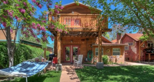 A charming wooden house with a balcony, surrounded by greenery and blooming flowers, featuring a hammock in the yard.