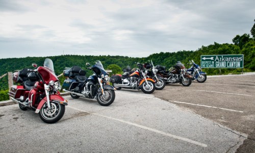 A row of motorcycles parked near a sign for the Arkansas Grand Canyon, surrounded by lush green hills under a cloudy sky.