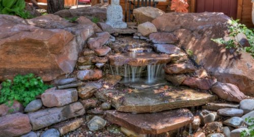 A serene stone waterfall surrounded by rocks and greenery, with a statue of a Buddha in the background.