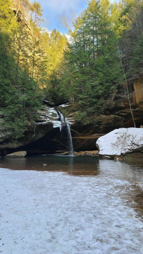 A serene winter scene featuring a waterfall cascading into a pool, surrounded by snow and lush green trees.