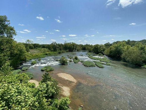 A serene river flows through a lush green landscape under a clear blue sky with scattered clouds.