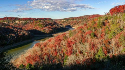 A scenic view of a river winding through vibrant autumn foliage in a hilly landscape under a partly cloudy sky.