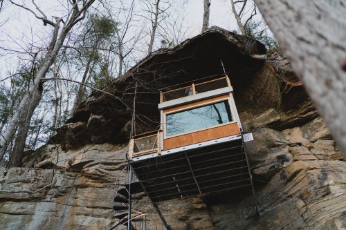 A modern glass cabin perched on a rocky cliff, surrounded by trees, with a spiral staircase leading up to it.