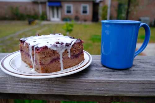 A slice of cake with icing on a plate next to a blue mug, set against a blurred outdoor background.