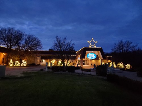 A building decorated with festive lights and a large star on top, surrounded by illuminated holiday figures at dusk.