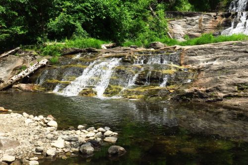 A serene waterfall cascades over rocks into a calm pool, surrounded by lush greenery and pebbled shoreline.