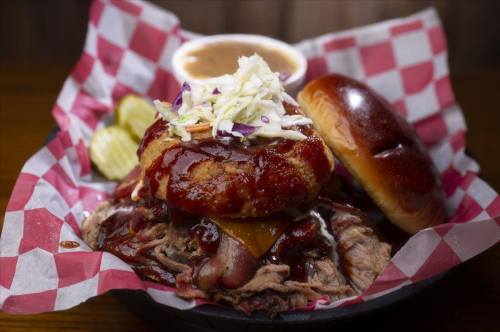 A plate of pulled pork topped with coleslaw and barbecue sauce, served with a bun, pickles, and a side of sauce.