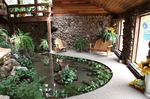 A cozy indoor space with a stone wall, two wooden chairs, and a small pond surrounded by lush greenery.