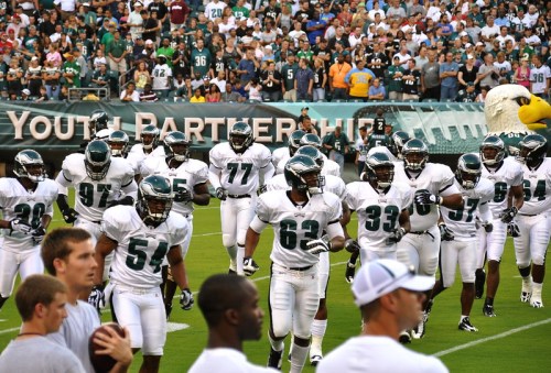 A group of football players in white uniforms runs onto the field, with a cheering crowd in the background.