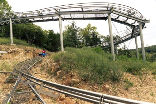 A person rides a mountain coaster on a winding track surrounded by greenery and rocky terrain.