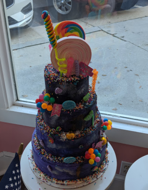 A colorful, multi-tiered cake decorated with candy, sprinkles, and a vibrant lollipop on top, set by a window.