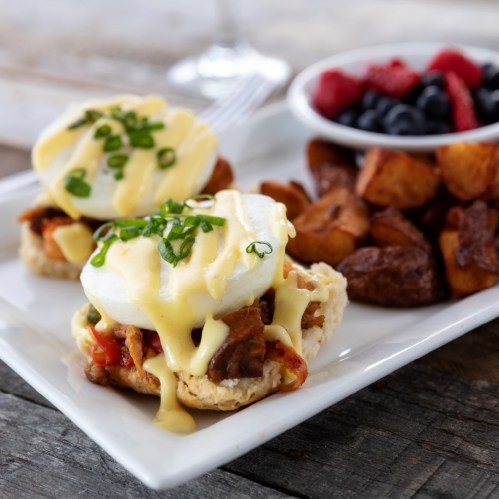 Two poached eggs on biscuits with hollandaise sauce, served with roasted potatoes and a side of mixed berries.