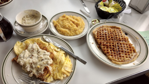 A table with a plate of fried chicken and gravy, a bowl of soup, a plate of grits, and a waffle.