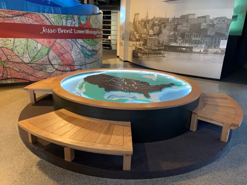 Interactive display featuring a 3D map of the Lower Mississippi River, surrounded by wooden seating.