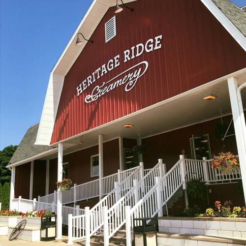 Red barn-style building with "Heritage Ridge Creamery" sign, white stairs, and colorful flower planters.