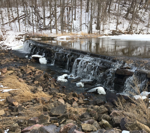 A frozen waterfall cascades over a stone dam, surrounded by snow-covered rocks and bare trees in a winter landscape.