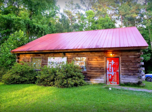 A rustic log cabin with a red metal roof, surrounded by greenery and featuring a weathered red door.