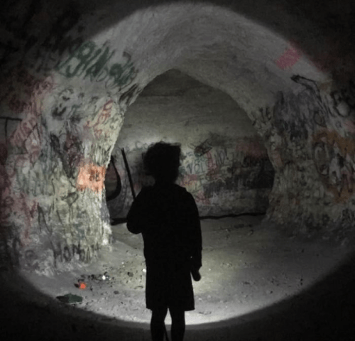A child stands in a dimly lit cave, holding a flashlight, surrounded by graffiti on the walls.