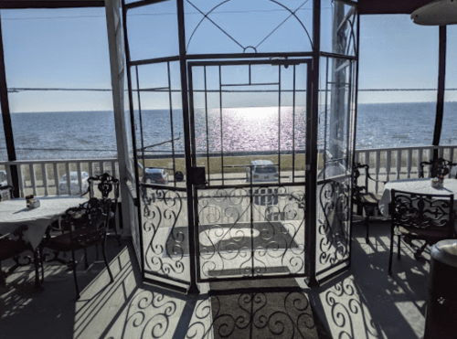 View from a restaurant with ornate doors opening to a sunny waterfront scene and tables set for dining.