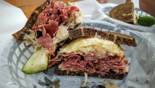 A close-up of a stacked pastrami sandwich with sauerkraut and melted cheese, served with a pickle on the side.
