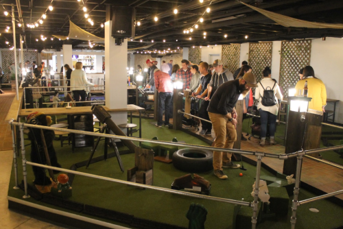 A lively indoor mini-golf course with people playing, string lights overhead, and various themed obstacles.