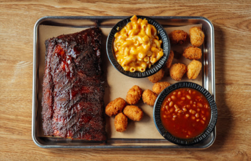 A metal tray with BBQ ribs, macaroni and cheese, baked beans, and crispy tater tots on a wooden table.