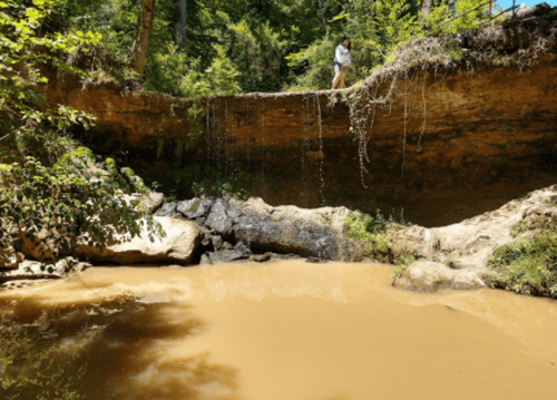 A person stands on a rocky ledge above a muddy creek, surrounded by lush greenery and cascading water.