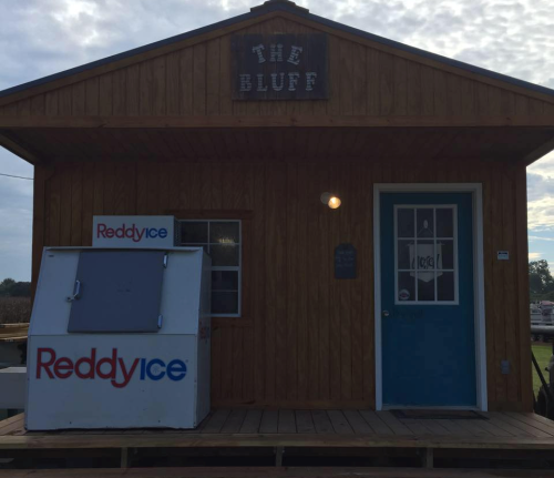 A wooden building with a sign reading "The Bluff" and a blue door, featuring a Reddy Ice vending machine outside.