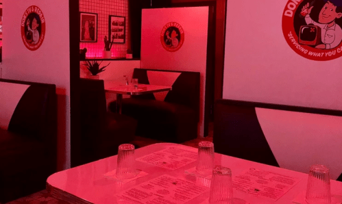 A retro diner interior with red lighting, featuring booths, tables, and menus on the tables.