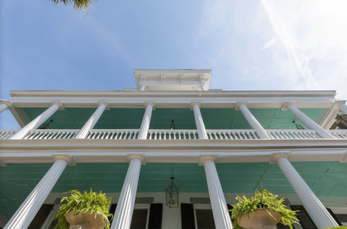A historic building with a turquoise ceiling, white columns, and hanging plants, viewed from below against a clear sky.