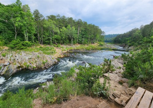 A scenic river flows through rocky terrain, surrounded by lush green trees under a cloudy sky.