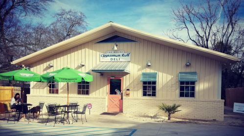 A light-colored deli building with a sign reading "Cinnamon Roll Deli," featuring outdoor seating and green umbrellas.